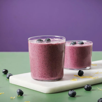 Two glasses filled with PACKD "get up and go" smoothie on a white board surrounded by loose blueberry's