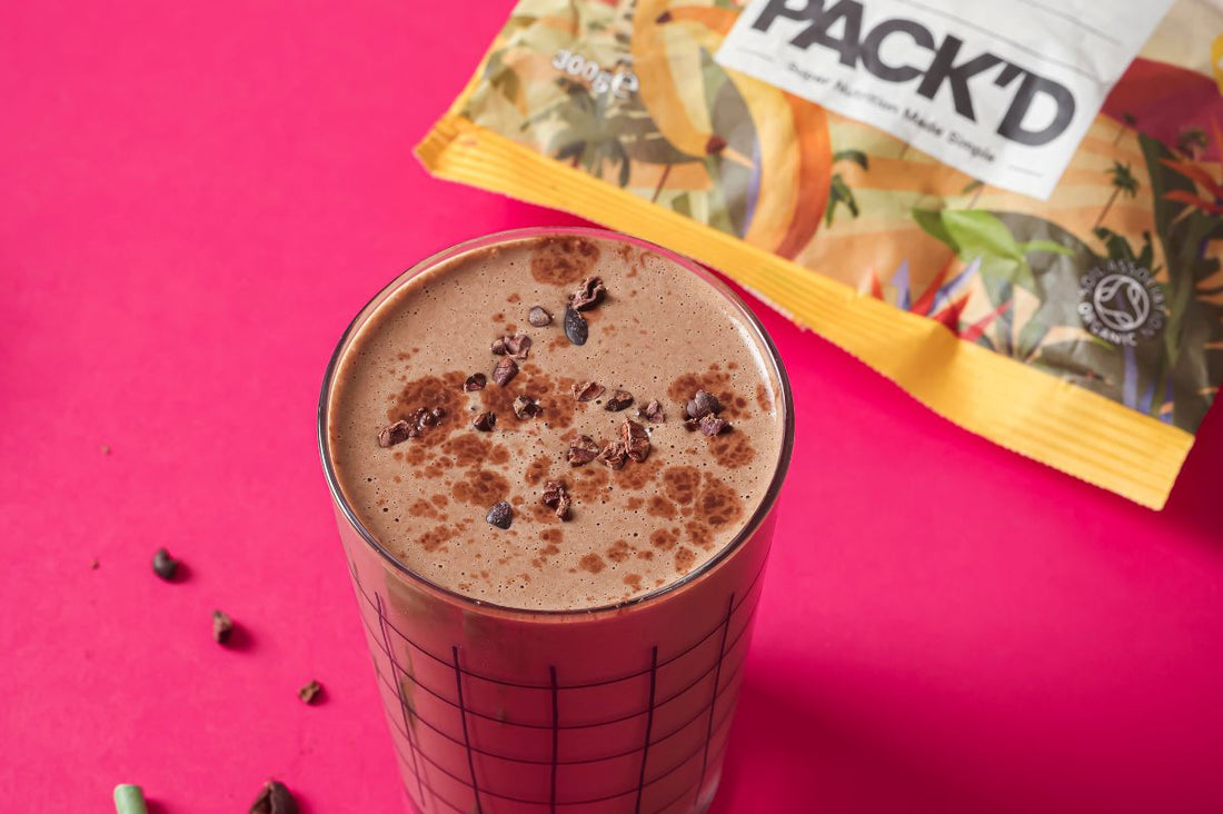 Glass filled with PACKD hormone balance smoothie on a red tablecloth next to a packet of PACKD frozen banana