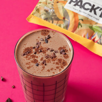 Glass filled with PACKD hormone balance smoothie on a red tablecloth next to a packet of PACKD frozen banana