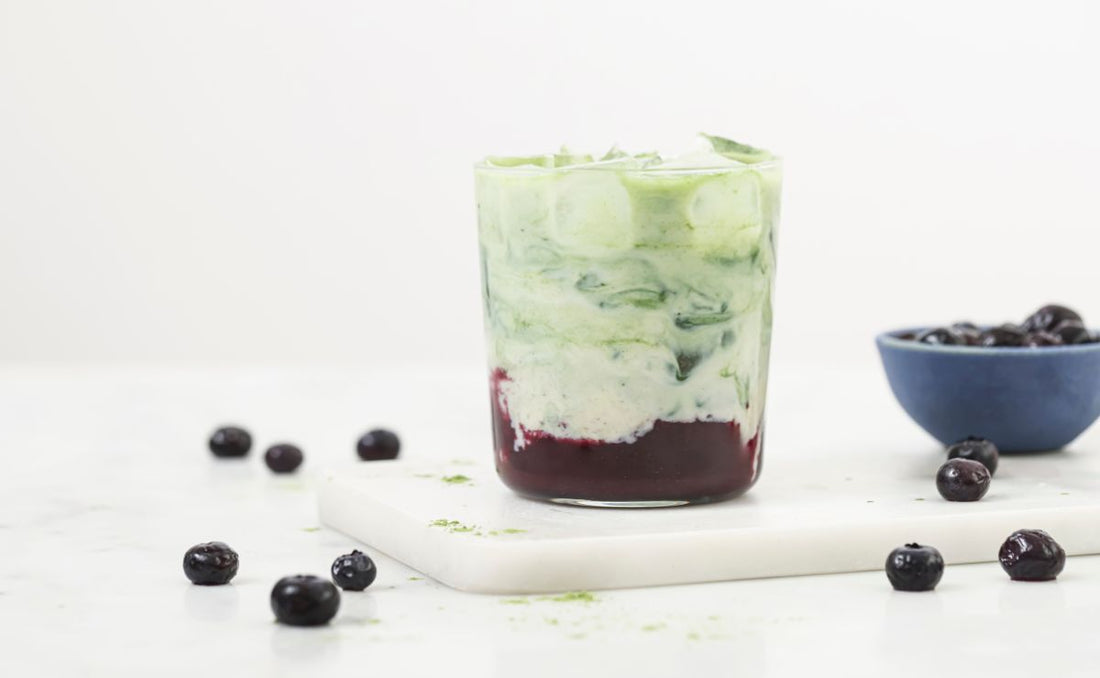 Blueberry iced matcha latte in a glass tumbler with a deep purple blueberry syrup layer beneath swirled green matcha and oat milk, surrounded by PACK'D Organic Frozen Blueberries on a marble board