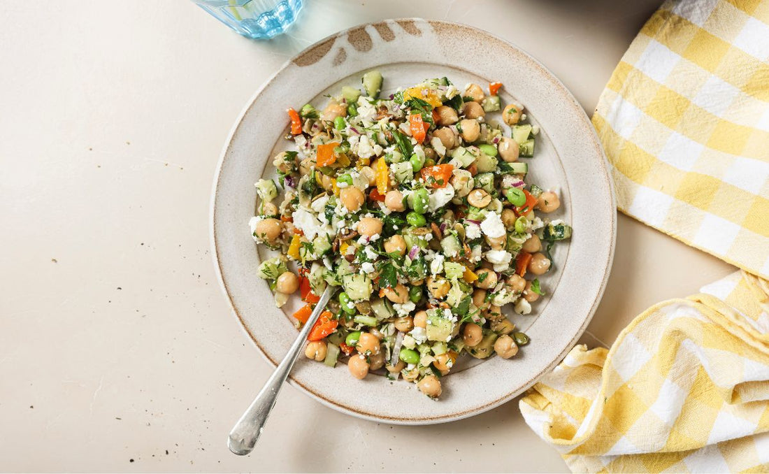 Overhead view of a stone bowl filled with chickpea and edamame crunch salad, with crumbled feta, diced cucumber, red onion, orange and yellow peppers, fresh dill and toasted pumpkin seeds, made with PACK'D Organic Edamame.