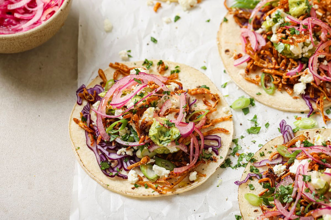 Three Crispy Chipotle Tofu Tacos served on parchment paper, filled with grated tofu and topped with PACK'D avocado guacamole, pink pickled onions, red cabbage, feta and fresh coriander.