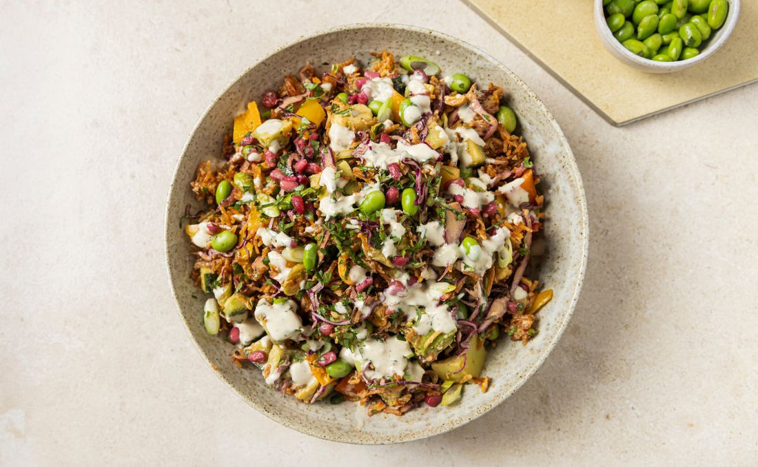 Crispy rice salad in a speckled ceramic bowl with PACK'D Organic Edamame, PACK'D Organic Avocado, PACK'D Organic Pomegranate, red cabbage and peppers, drizzled with ginger tahini dressing, beside a small bowl of edamame
