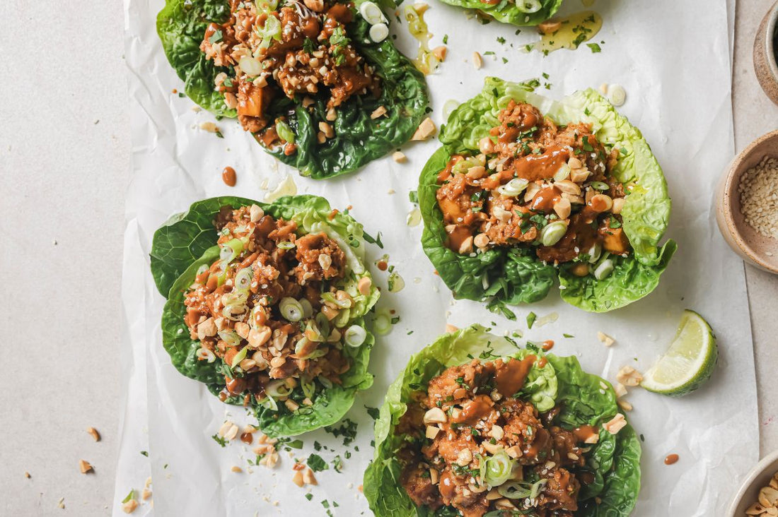 Overhead view of four Firecracker Tofu and Cauliflower Lettuce Wraps on parchment paper, filled with crispy tofu in peanut sauce and topped with chopped peanuts, spring onions, and coriander.