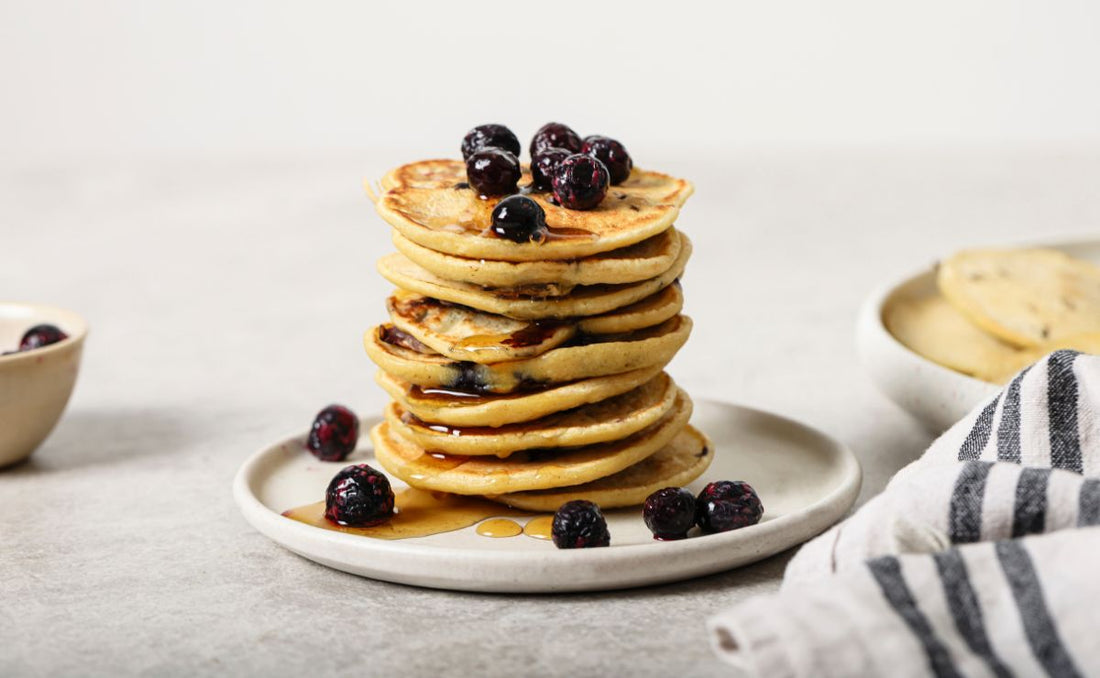 Stack of fluffy blueberry pancakes topped with PACK'D Organic Blueberries and maple syrup on a white plate