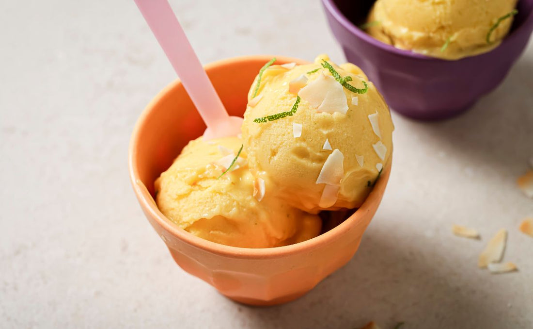 Two scoops of golden mango, coconut and lime ice cream in a terracotta bowl with a pink spoon, topped with toasted coconut flakes and lime zest, made with PACK'D Organic Mango.