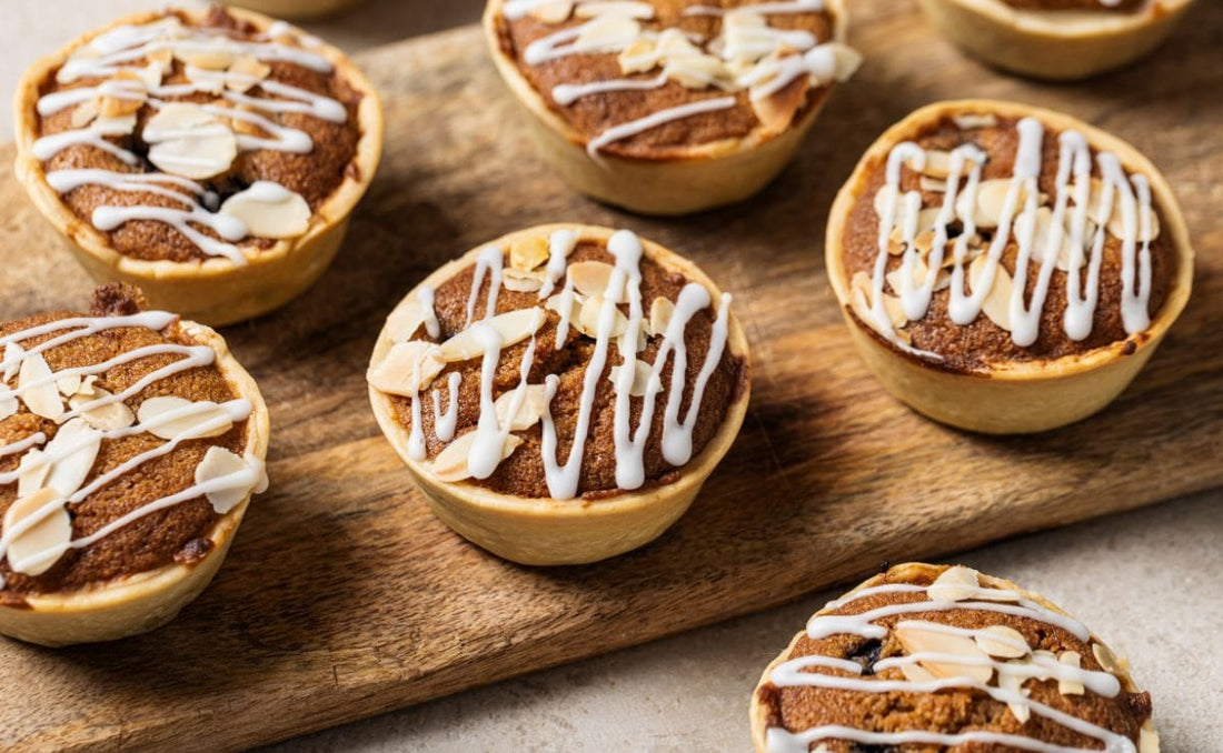 Mini blueberry Bakewell pies in shortcrust pastry cases, topped with flaked almonds and a white icing drizzle on a wooden board, made with PACK'D Organic Frozen Blueberries