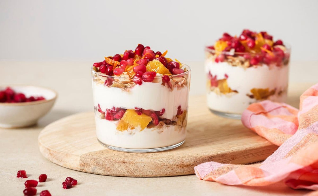 Two glass jars layered with creamy Greek yoghurt, crunchy granola, diced orange segments, and bright red PACK'D Organic Pomegranate Arils.
