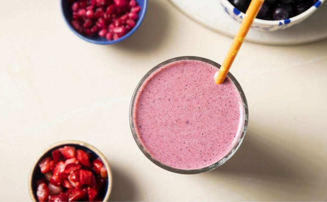 Overhead view of a pink pomegranate and berry smoothie in a glass with an orange paper straw, surrounded by bowls of pomegranate seeds, chopped strawberries and PACK'D Organic Blueberries.