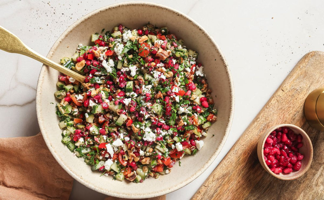 Overhead view of a large bowl filled with a vibrant salad of PACK'D Organic Pomegranate Arils, toasted walnuts, diced cucumber, tomatoes, and crumbled feta.