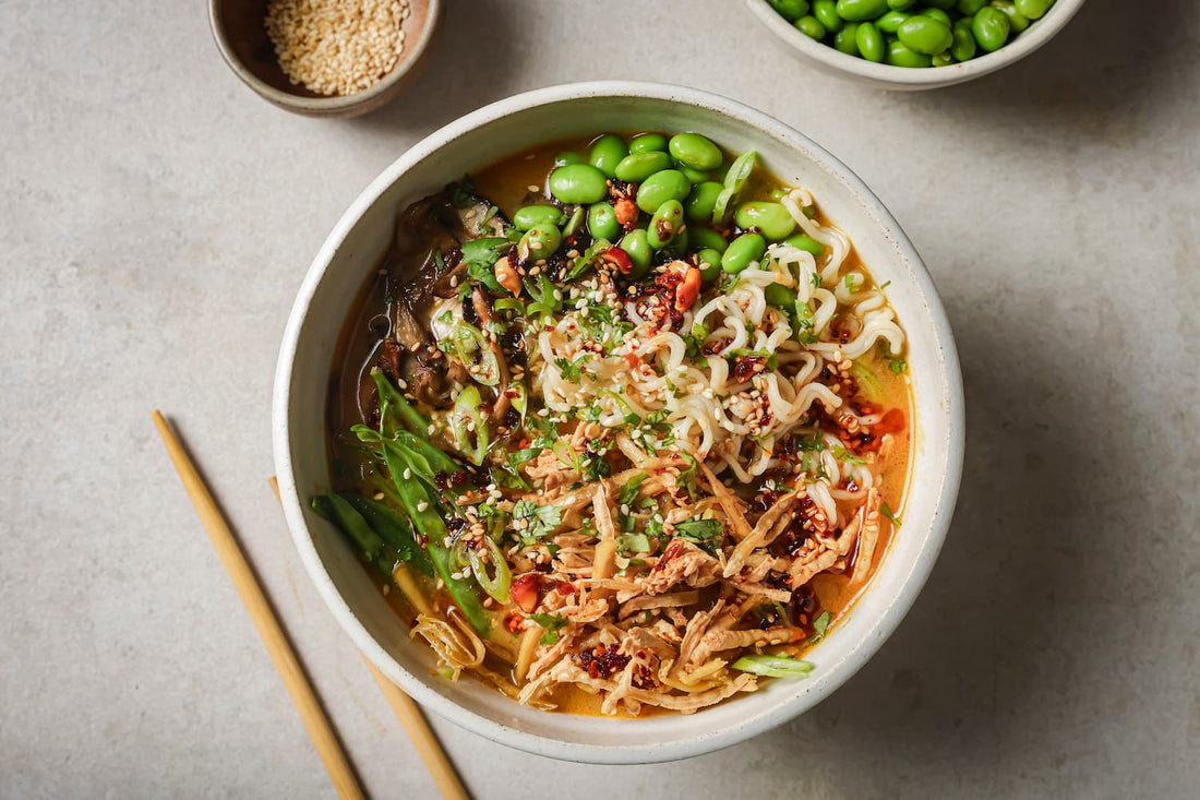 Overhead view of a bowl of Spicy Peanut Ramen filled with noodles, crispy grated tofu, glazed oyster mushrooms, bright green PACK'D edamame beans and chilli crisp.