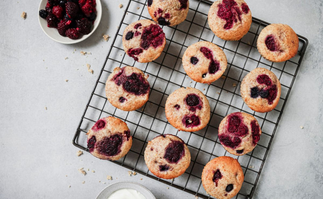 Twelve summer berry yoghurt muffins cooling on a wire rack, studded with PACK'D Organic Summer Berries, with a bowl of frozen berries and a dish of Greek yoghurt alongside.