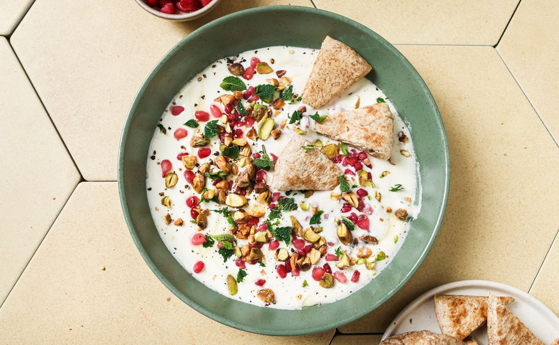 Whipped feta dip topped with PACK'D Organic Pomegranate Seeds, chopped pistachios and torn mint in a green ceramic bowl, served with toasted pitta triangles on a beige tiled surface